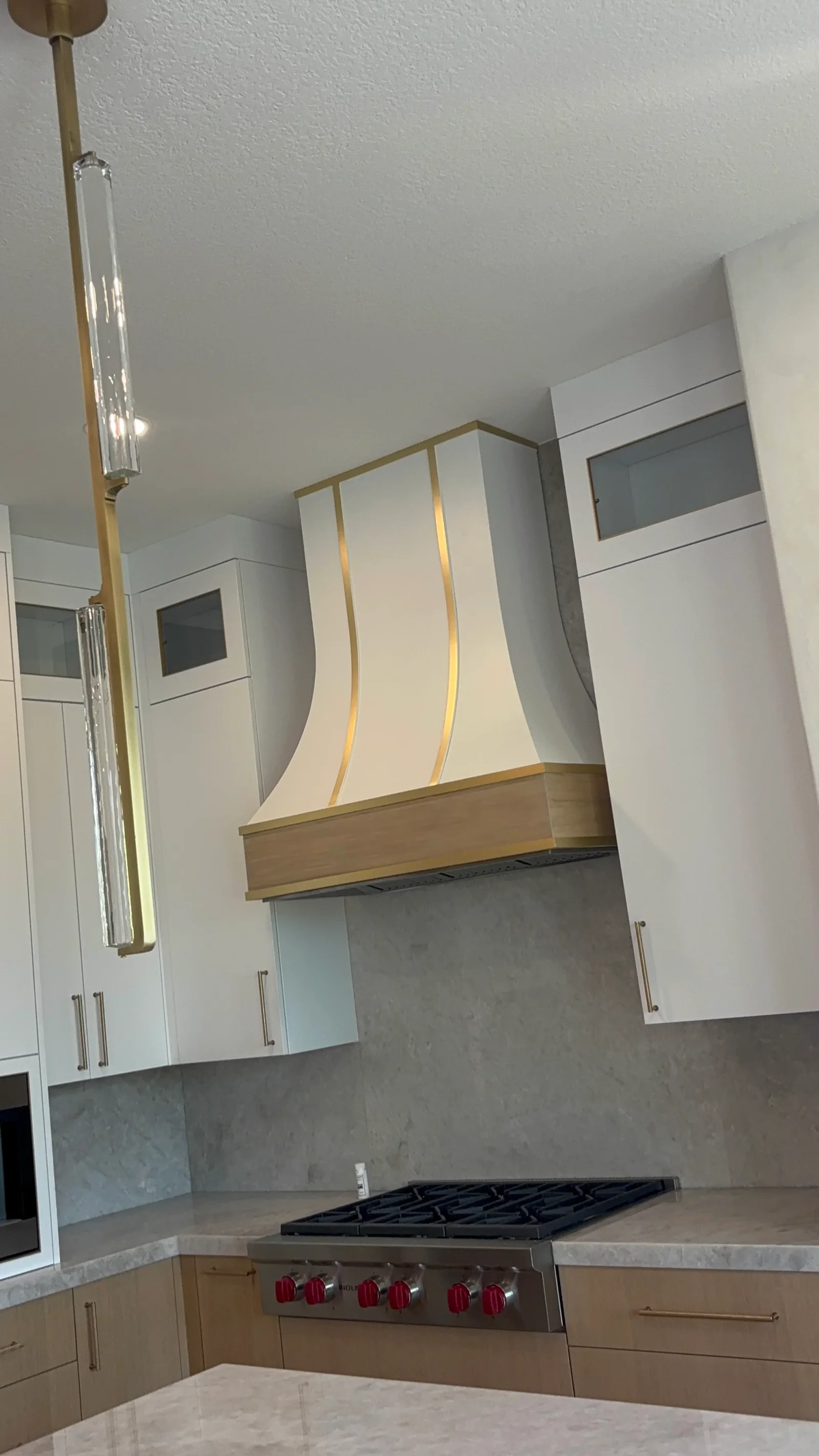 White painted kitchen cabinets with curved cream and brass range hood, gray backsplash, and textured ceiling